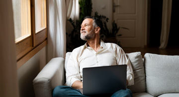 Man sitting in a relaxed position on his sofa at home, with a laptop on his lap.