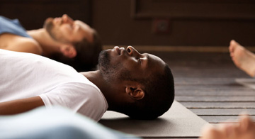 Male lying down on a yoga mat, with his eyes closed, in the middle of a yoga class