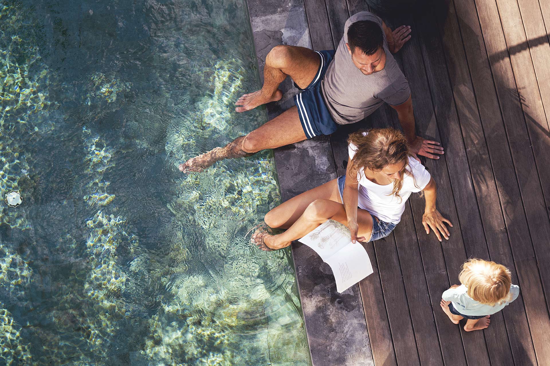 , image shows a family sitting on wooden decking with their feet in the water, reading to a young child