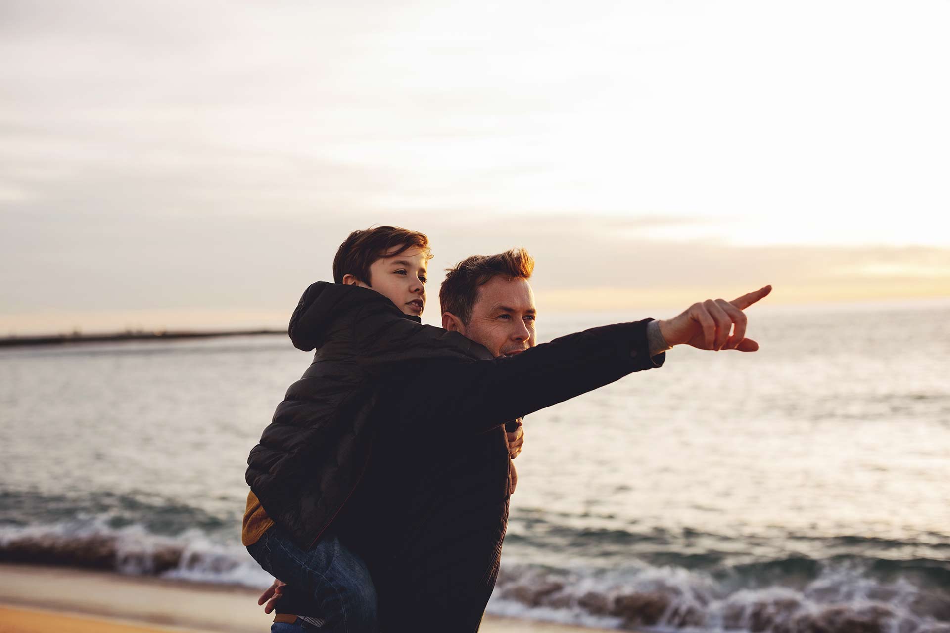 , image shows a father carrying a child on his back at a beach, pointing into the distance